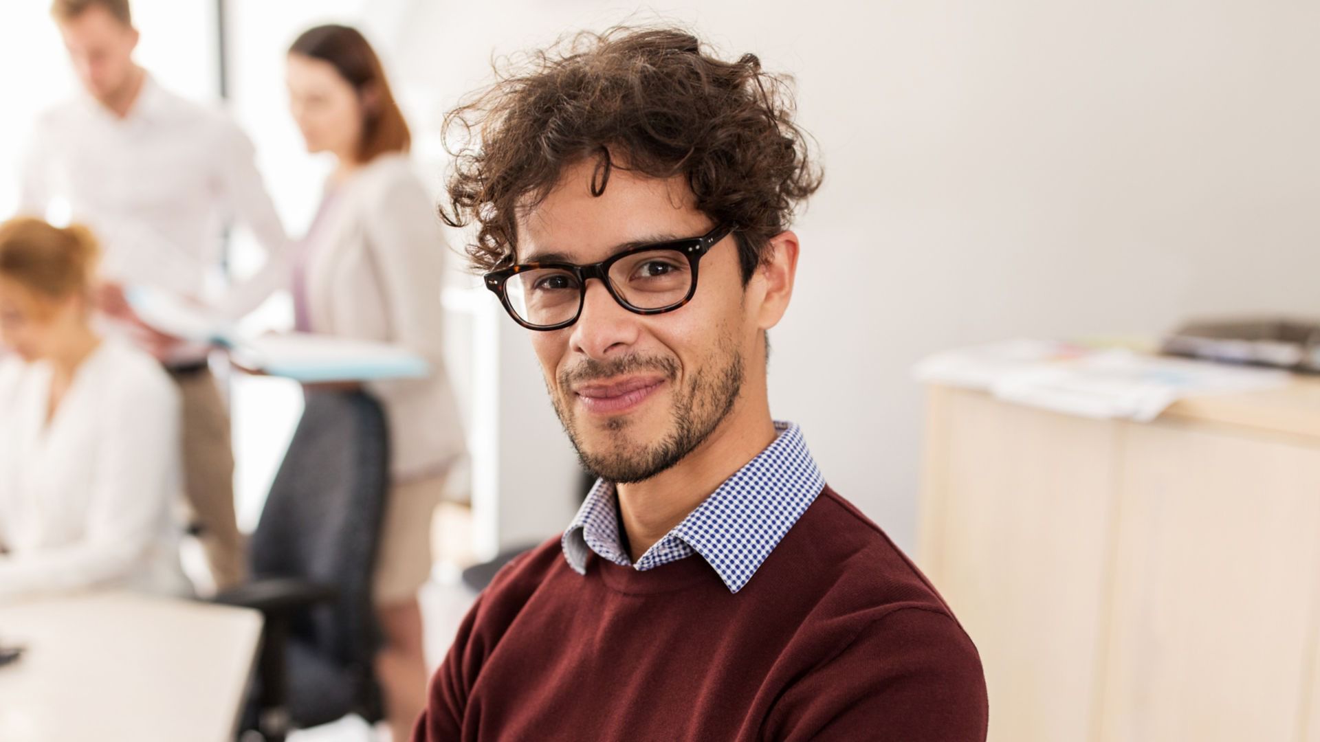 Man looking into the camera wearing dark rimmed glasses and maroon sweater with collared shirt.