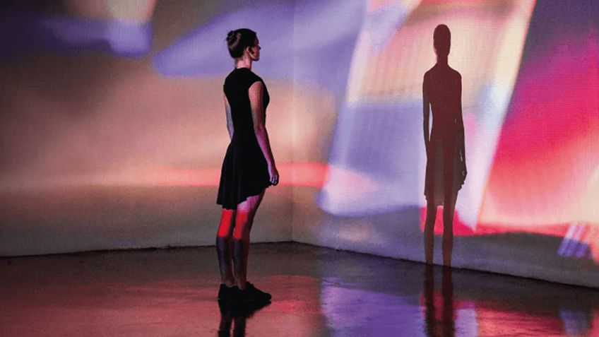 Person in a black dress stands in a colorful digital art installation with vibrant projections on walls and a reflective floor.