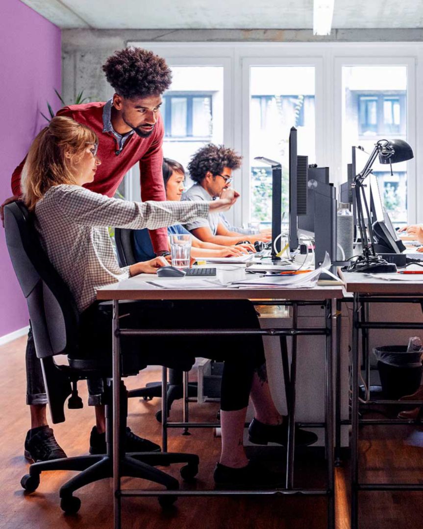 A colleague helping another colleague who is seated along a row of people working on their computers.