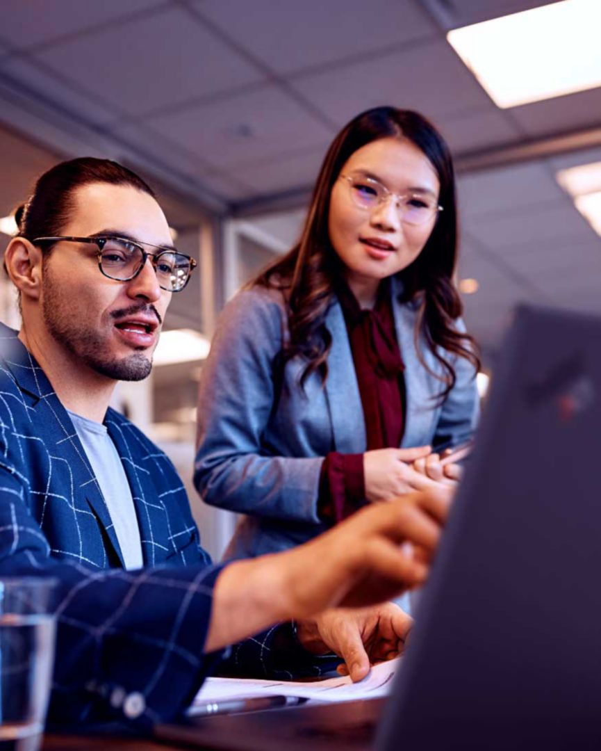 A female colleague leaning over the shoulder of her male colleague looking at his laptop screen.