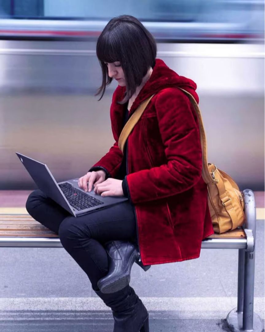 A woman seated on a bench using her laptop as a train passes behind her.