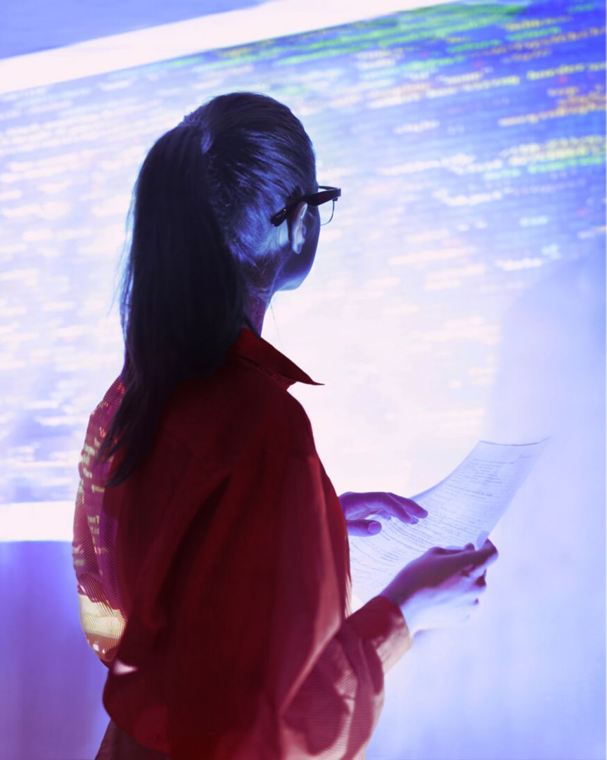 A woman holding a sheet of paper and looking at data projected on the wall.