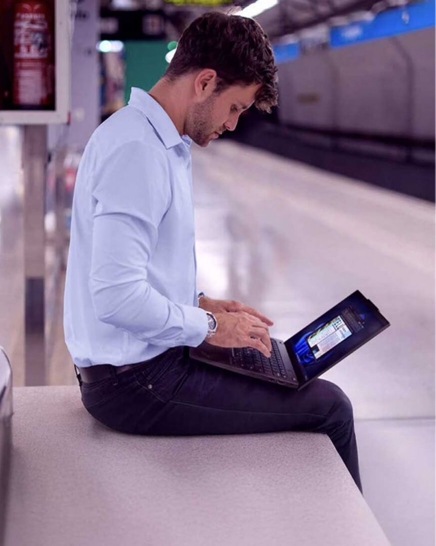 A man seated on a bench in a train station using his laptop.