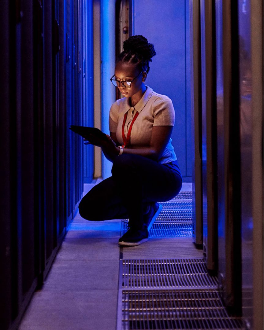 A woman kneeling on the ground in a dark server room using a laptop.