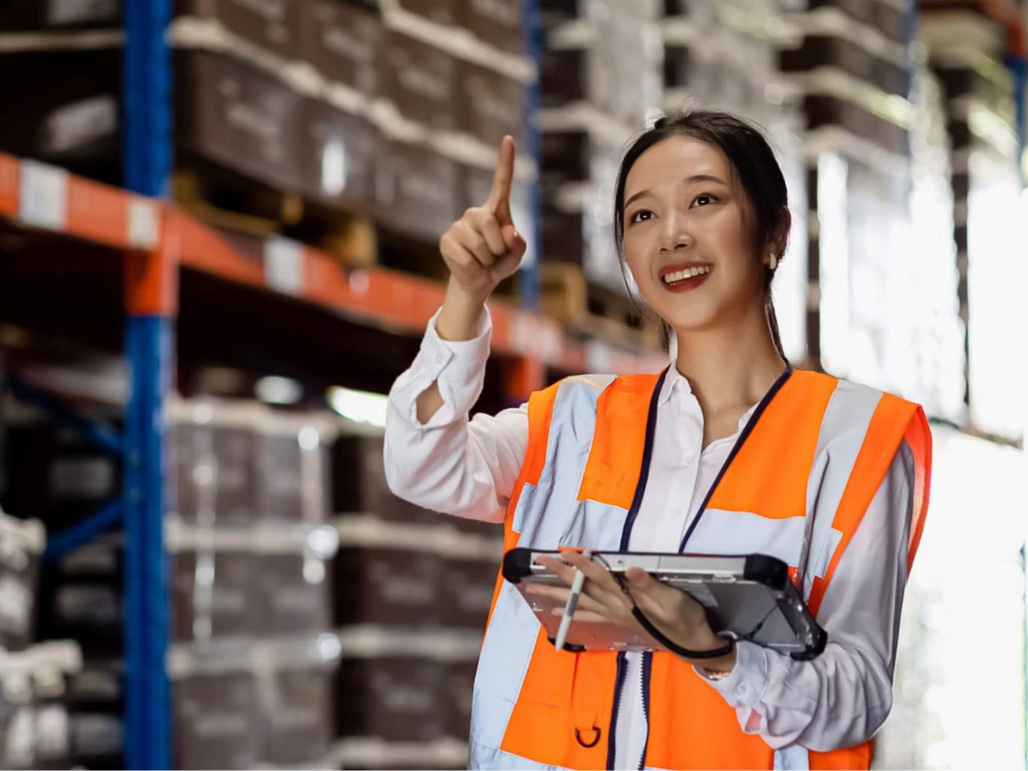 A woman wearing an orange safety vest and holding a tablet pointing in a warehouse.