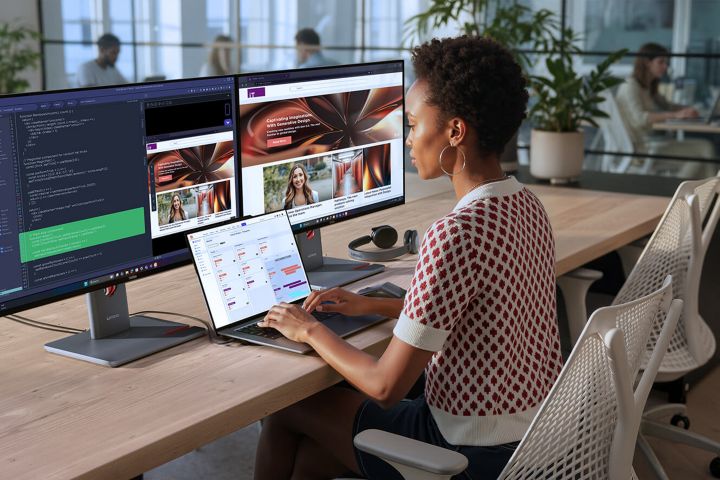 A woman typing on a laptop with two monitors behind it on her desk.