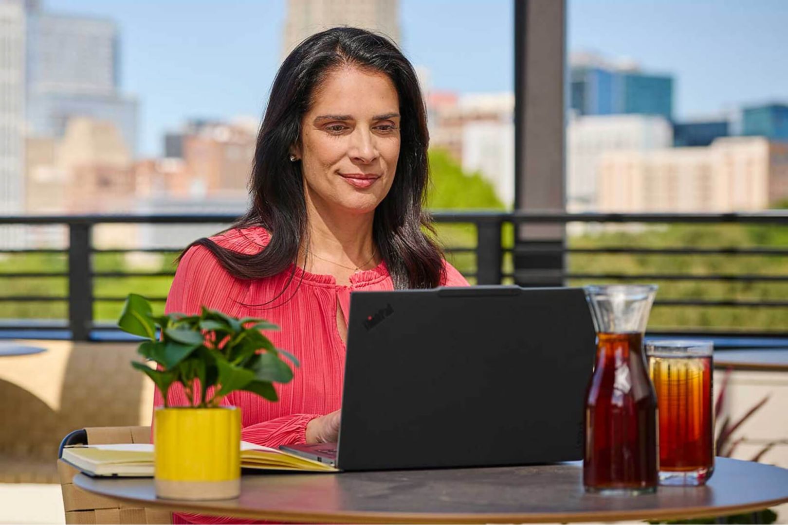 Una mujer trabajando en su laptop desde una mesa al aire libre y disfrutando de té helado.