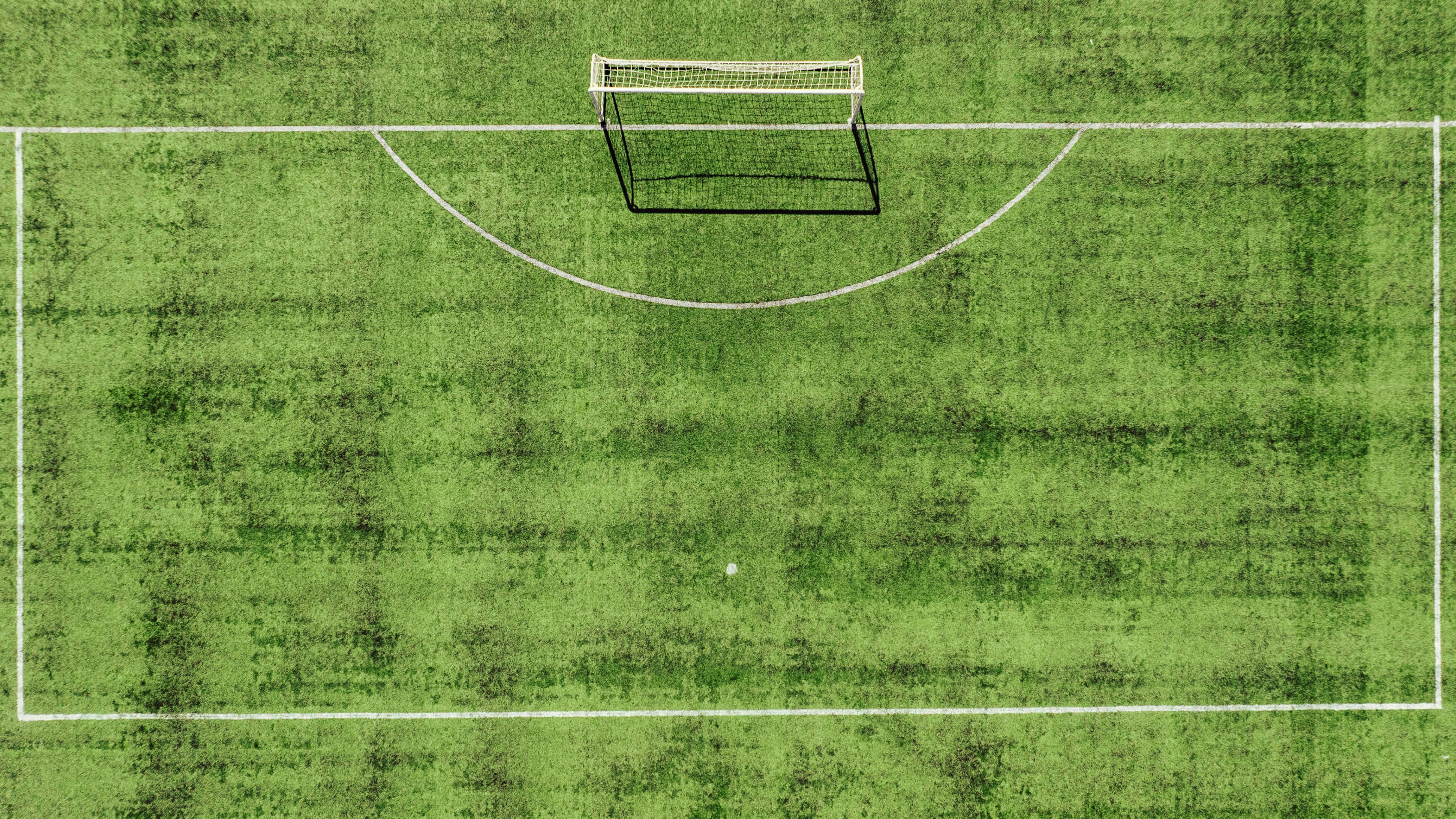 Aerial view of an empty green football pitch, showing a white goal, penalty area markings, and the goal’s long shadow.