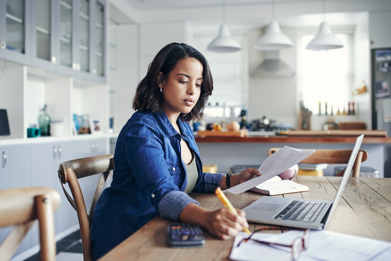 Shot of a young woman using a laptop and  going through paperwork while working from home