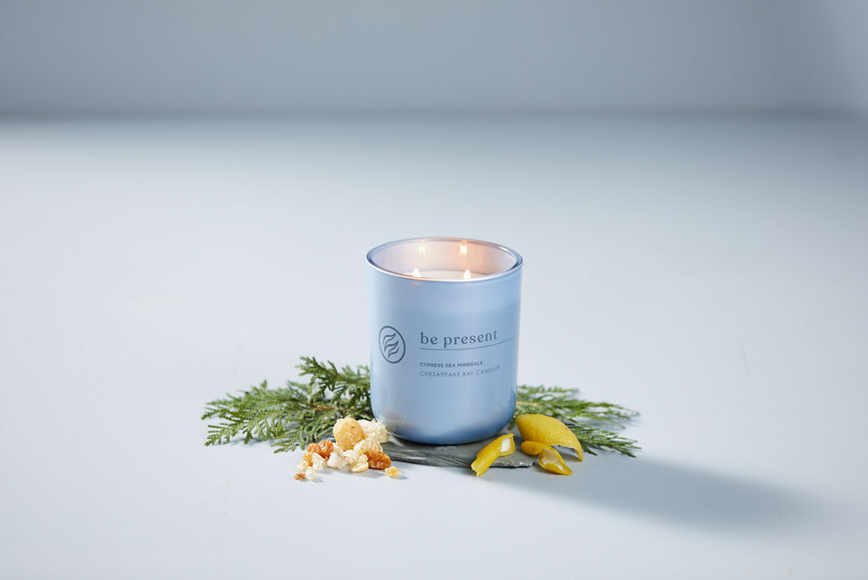 chesapeake bay candle intentions collection be present cypress sea minerals medium two wick jar candle surrounded by lemon pieces and a cypress branch in a pastel blue backdrop
