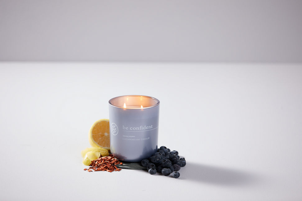 chesapeake bay candle intentions collection be confident indigo poppy medium two wick jar candle surrounded by blueberries and lemon slices in a pastel purple backdrop