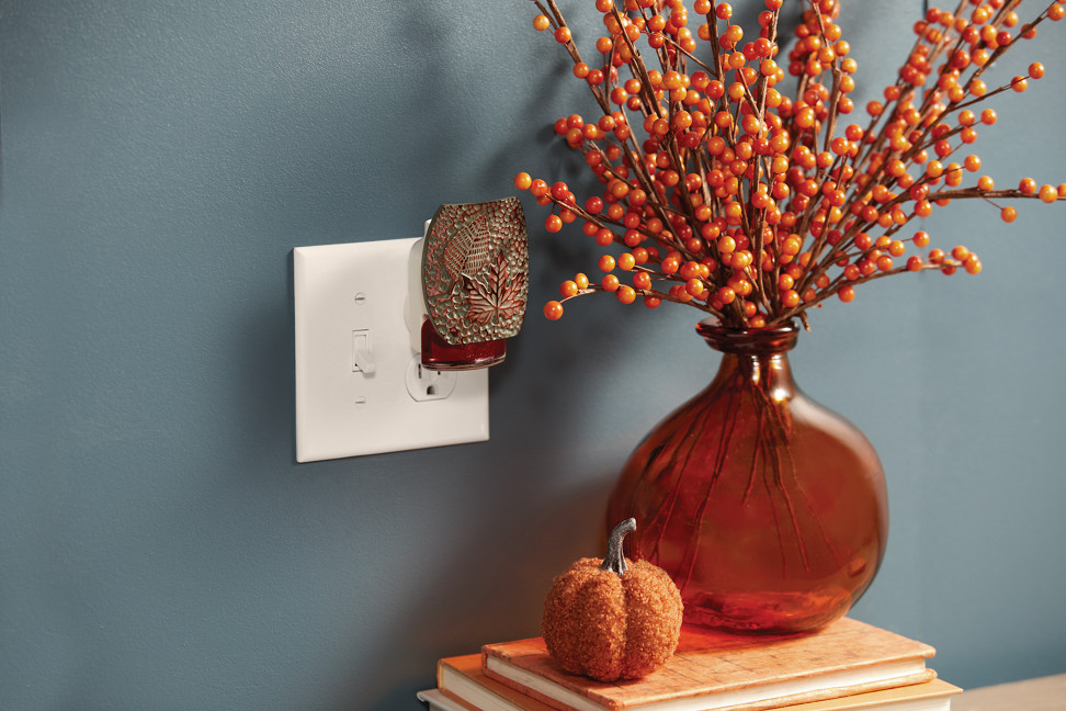 hammered leaves scentplug diffuser plugged into a wall with a scentplug refill above orange books, an orange decorative pumpkin, and an orange decorative plant in an orange vase