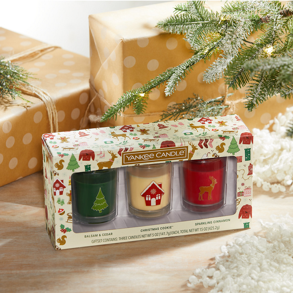 three 5 ounce candles in a gift set featuring the fragrances of Balsam & Cedar, Christmas Cookie, and Sparkling Cinnamon on a table with wrapped presents