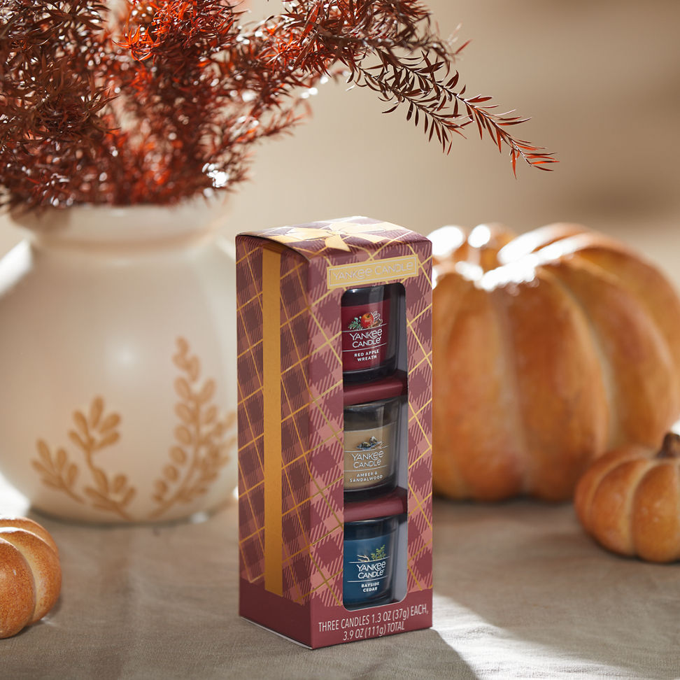 Sweater Weather Minis Gift Set featuring a Red Apple Wreath Yankee Candle Mini, an Amber & Sandalwood Yankee Candle Mini, and a Bayside Cedar Yankee Candle Mini on a table with pumpkins and flowers in a white vase