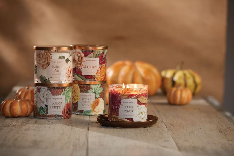 rainy day 3-Wick candle, macintosh 3-Wick candle, spiced pumpkin 3-Wick candle, pumpkin banana scone 3-Wick candle, and an autumn leaves 3-Wick candle on a wood table with pumpkins