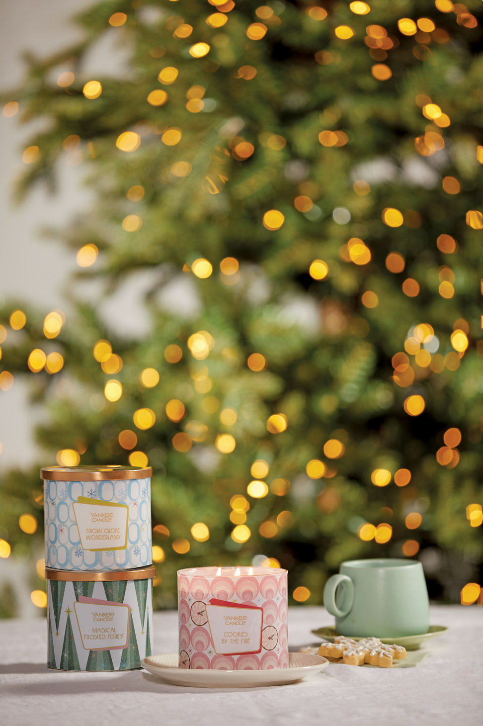 a snow globe wonderland 3-Wick candle, a magical frosted forest 3-Wick candle, and a cookies by the fire 3-Wick candle next to a green mug and a snowflake cookie on a white table with a sparkling tree in the background