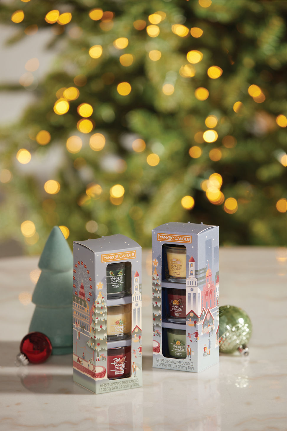 a nostalgic christmas classics minis gift set featuring a balsam & cedar yankee candle® mini, a christmas cookie™ yankee candle® mini, and a sparkling cinnamon yankee candle® mini and a holiday party minis gift set including a vanilla cupcake yankee candle® mini, a holiday zest yankee candle® mini, and a sage & citrus yankee candle® mini on a table with a green and red ornament and a decorative tree