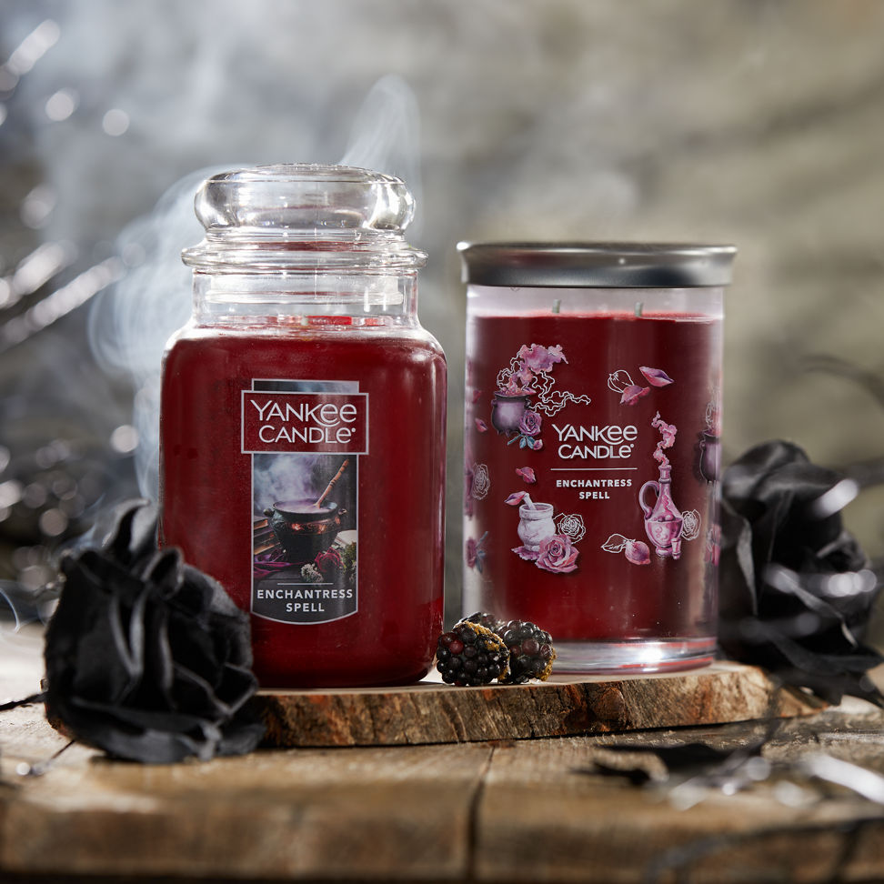 Enchantress Spell Original Large Jar Candle and Enchantress Spell Signature Large Tumbler Candle next to two black roses and blackberries on a piece of wood with smoke in the background