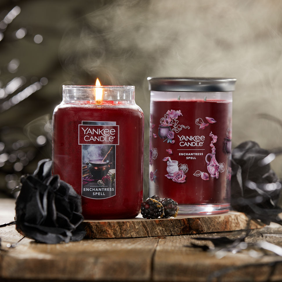 Enchantress Spell Original Large Jar Candle and Enchantress Spell Signature Large Tumbler Candle next to two black roses and blackberries on a piece of wood with smoke in the background