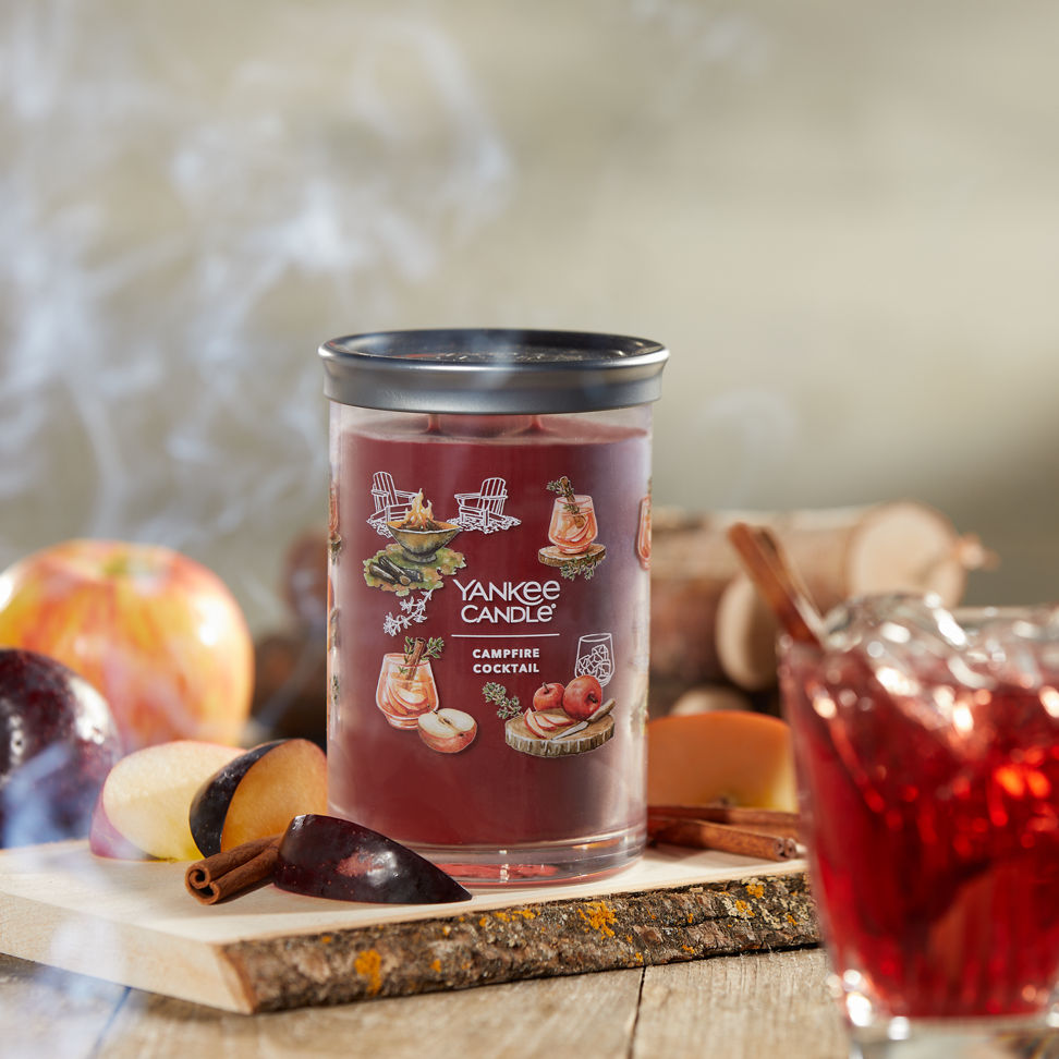 Cottage Getaway Collection Campfire Cocktail Signature Large Tumbler Candle on a piece of wood next to apple slices and a drink