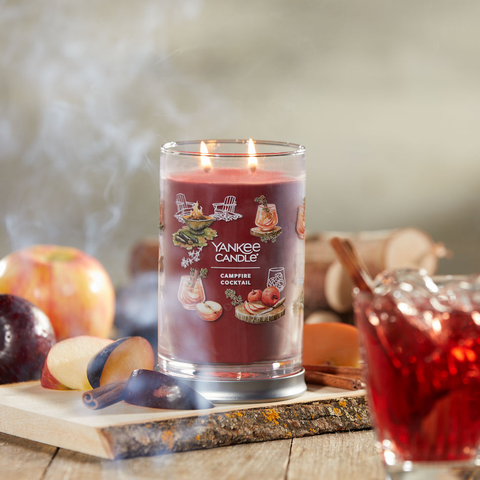 Cottage Getaway Collection Campfire Cocktail Signature Large Tumbler Candle on a piece of wood with slices of fruit and cinnamon sticks next to a mixed drink