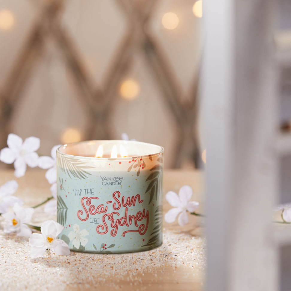 tis the sea-sun in sydney three wick candle on table with white flowers and sand