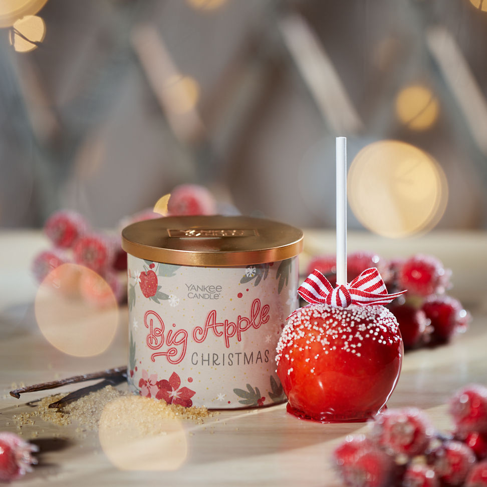 big apple christmas three wick next to a red candied apple among red berries vanilla beans and sugar with lights all around