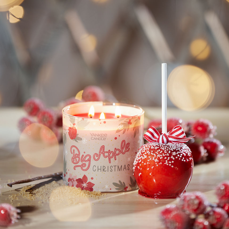 big apple christmas three wick next to a red candied apple among red berries, vanilla beans, and sugar with lights all around
