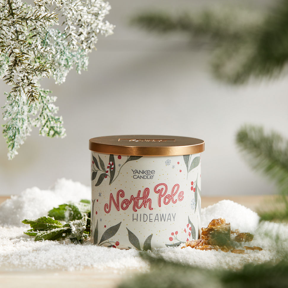 north pole hideaway three wick candle on surface with snow, green leaves, amber, and evergreen twigs