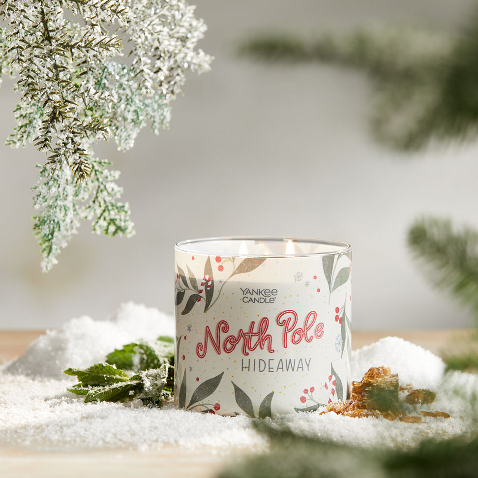north pole hideaway three wick candle on surface with snow, green leaves, amber, and evergreen twigs