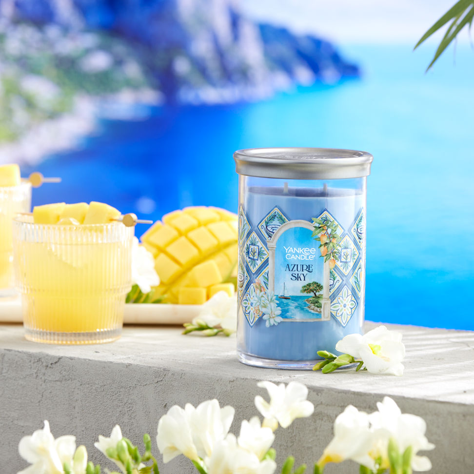 Hello, Italy! Collection Azure Sky Signature Large Tumbler Candle next to two mixed drinks, sliced fruit, and white flowers