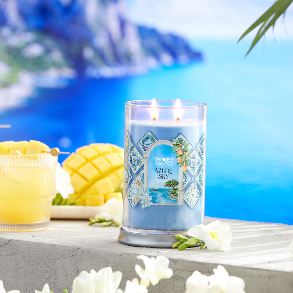 Hello, Italy! Collection Azure Sky Signature Large Tumbler Candle next to a mixed drink, sliced fruit, and white flowers