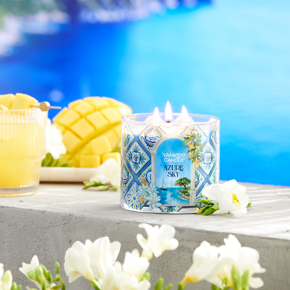 Hello, Italy! Collection Azure Sky 3-Wick Candle next to a mixed drink, sliced fruit, and white flowers