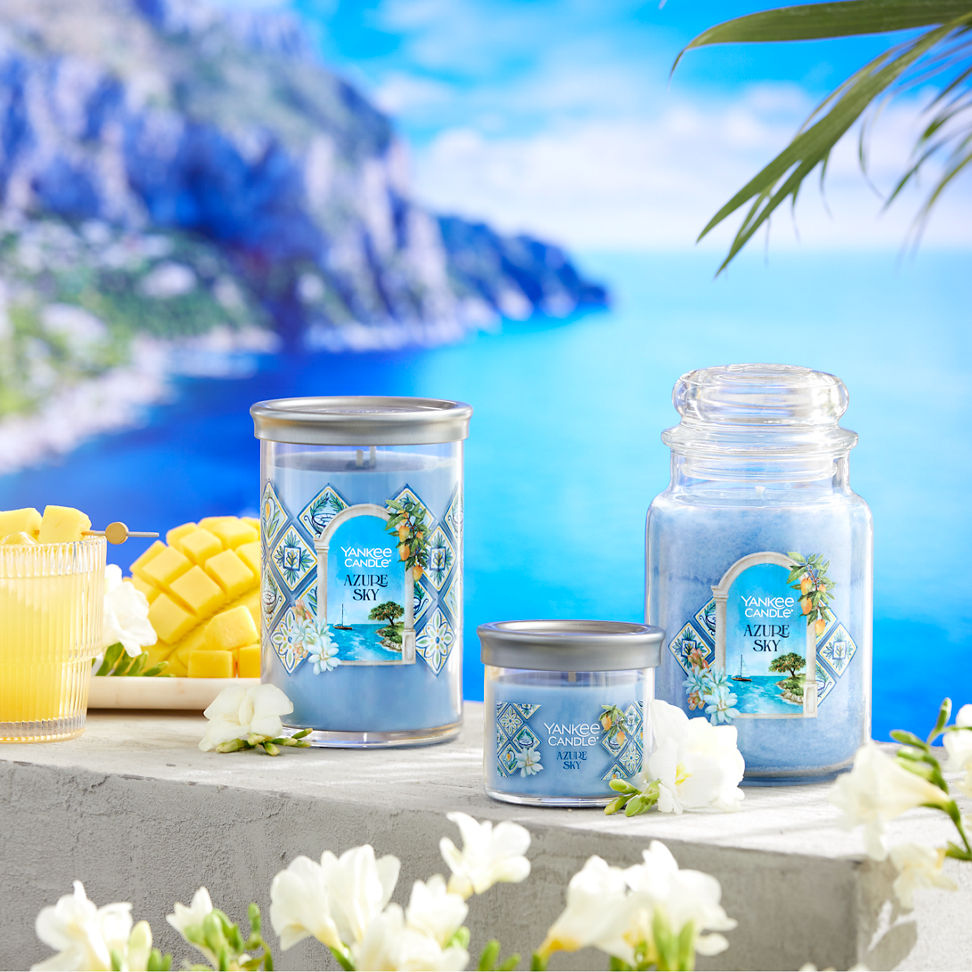Hello, Italy! Collection Azure Sky Signature Large Tumbler Candle, Azure Sky Signature Small Tumbler Candle, and Azure Sky Original Large Jar Candle on a concrete surface with sliced fruit, white flowers, and a mixed drink overlooking the water