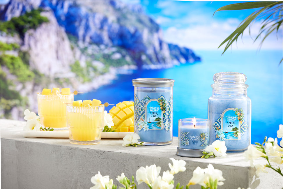 Hello, Italy! Collection Azure Sky Signature Large Tumbler Candle, Azure Sky Signature Small Tumbler Candle, and Azure Sky Original Large Jar Candle on a concrete surface next to sliced fruit, two mixed drinks, and white flowers