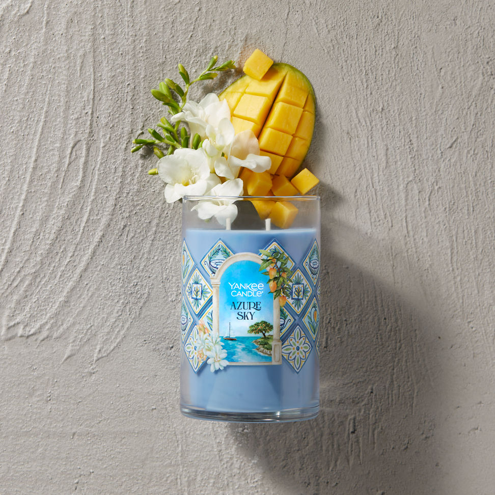 Hello, Italy! Collection Azure Sky Signature Large Tumbler Candle laying on a concrete surface with sliced mango and white flowers coming from the top of the candle
