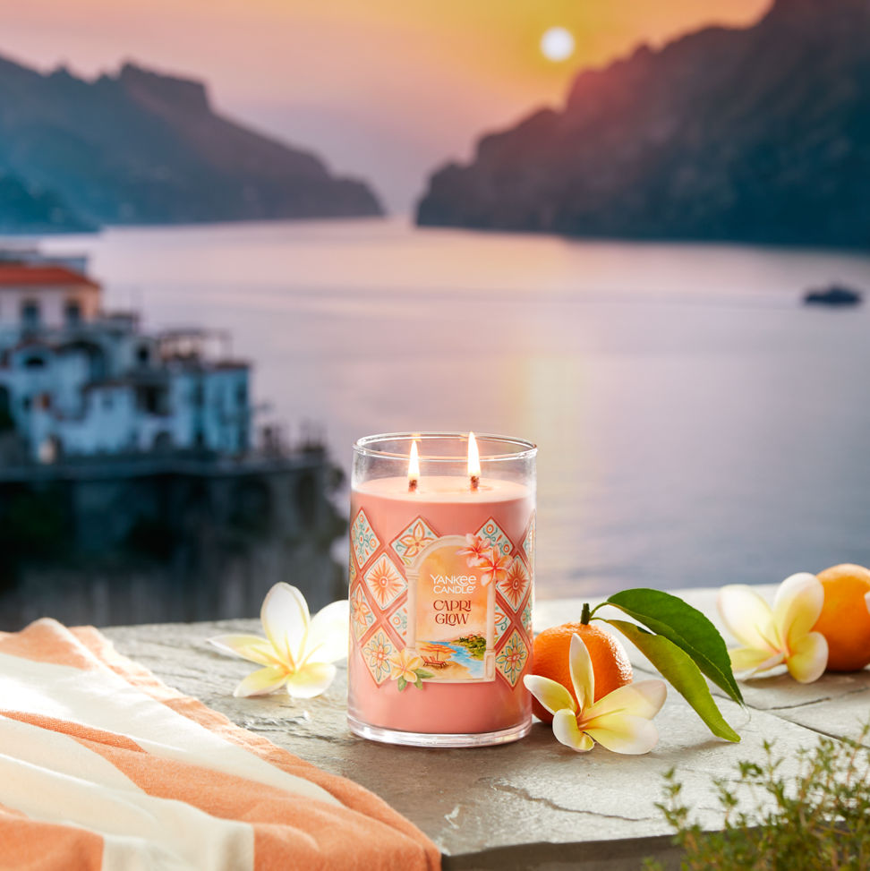 Hello, Italy! Collection Capri Glow Signature Large Tumbler Candle on a concrete step with flowers and fruit overlooking a sunset and water