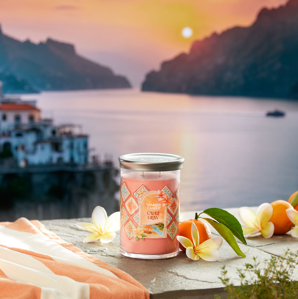 Hello, Italy! Collection Capri Glow Signature Large Tumbler Candle on a concrete step with flowers and fruit overlooking a sunset and water