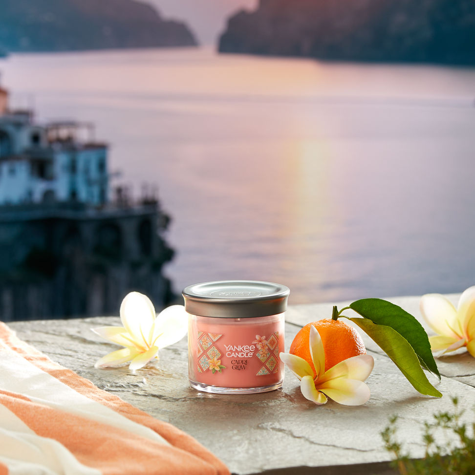 Hello, Italy! Collection Capri Glow Signature Small Tumbler Candle on a concrete step with flowers and fruit overlooking a sunset and water