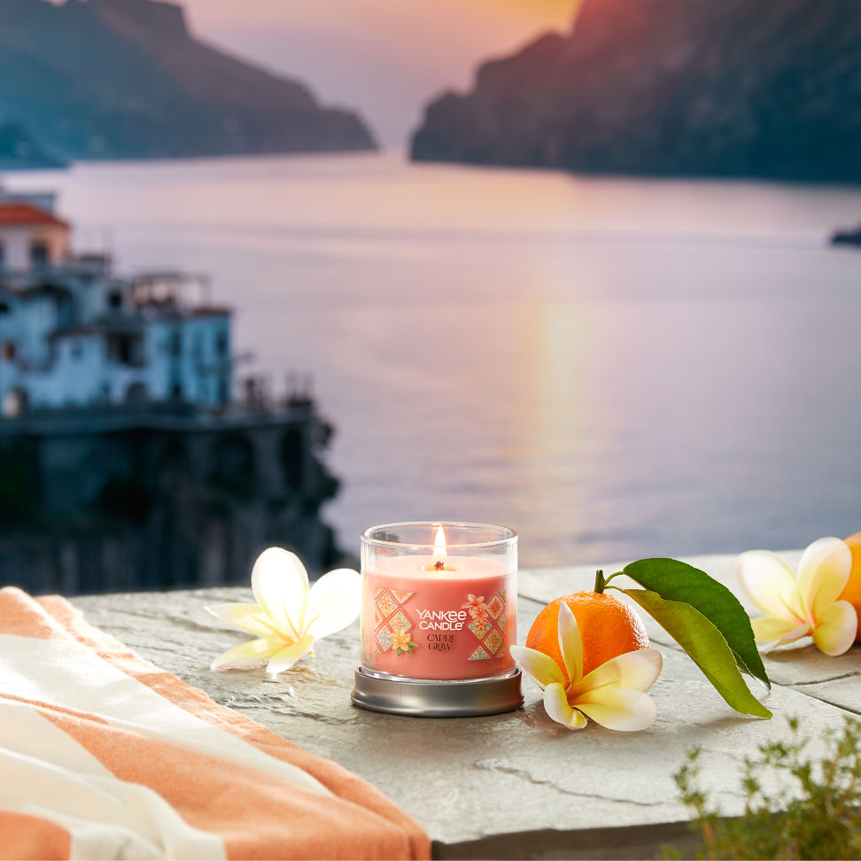 Hello, Italy! Collection Capri Glow Signature Small Tumbler Candle on a concrete step with flowers and fruit overlooking water and a sunset