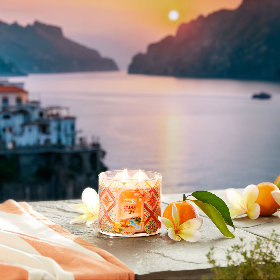 Hello, Italy! Collection Capri Glow 3-Wick Candle on a concrete surface with flowers and fruit overlooking water and a sunset