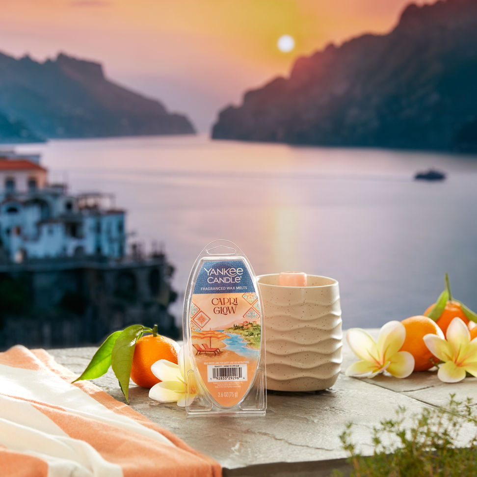 Hello, Italy! Collection Capri Glow Wax Melts (6-Pack) and a Sandstone Wax Warmer on a concrete surface with flowers and fruit overlooking water and a sunset