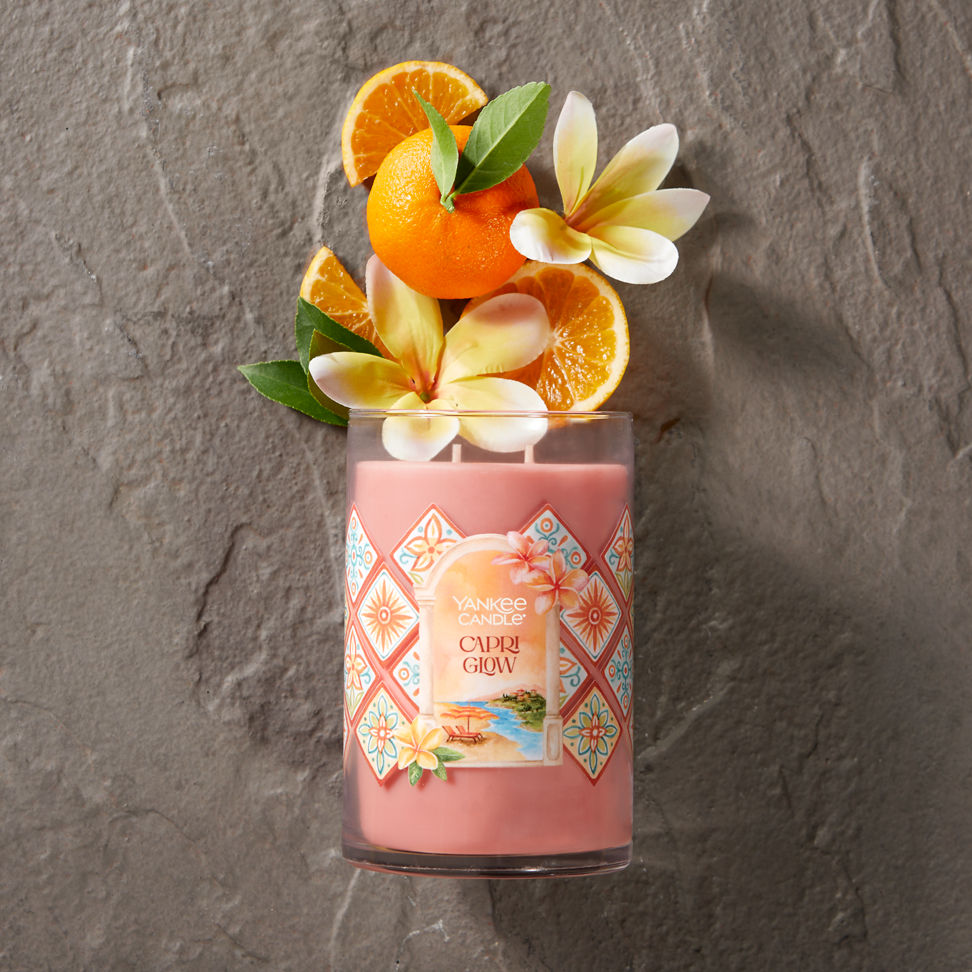 Hello, Italy! Collection Capri Glow Signature Large Tumbler Candle laying on a concrete surface with slices of fruit and flowers coming from the top of the candle