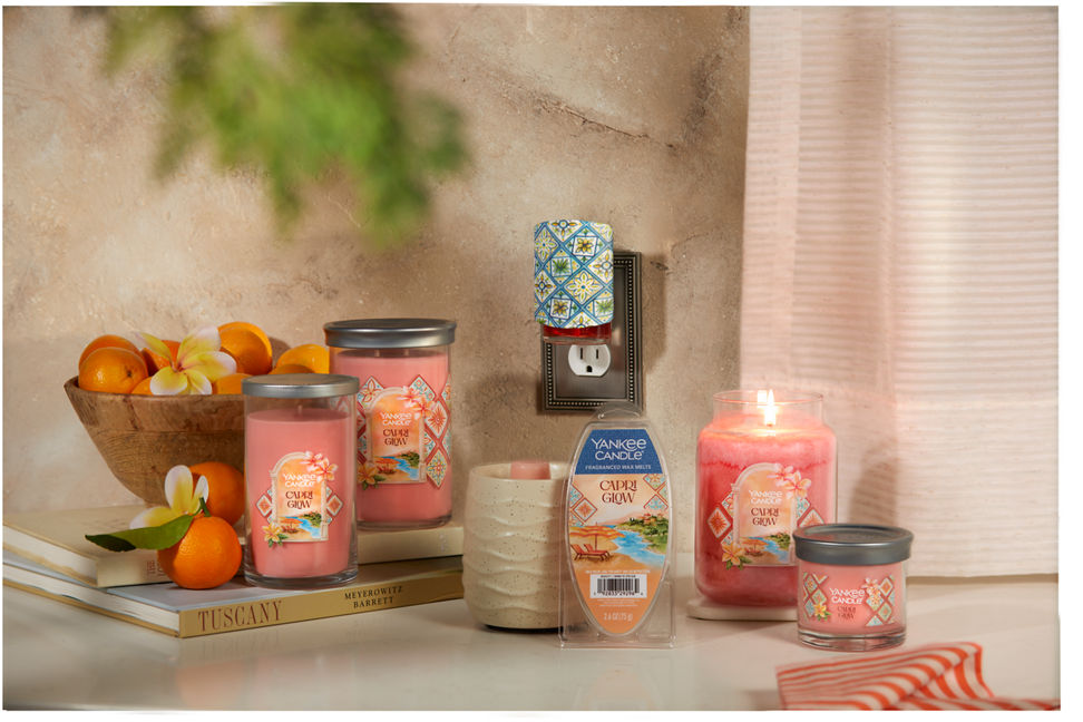 Hello, Italy! Collection Capri Glow Signature Medium Pillar Candle, Capri Glow Signature Large Tumbler Candle, a Sandstone Wax Warmer, Capri Glow Wax Melts 6-Pack, Hello, Italy! ScentPlug Diffuser with a light, Capri Glow Original Large Jar Candle, and Capri Glow Signature Small Tumbler Candle on a table with two books and a bowl of small tangerines and yellow flowers