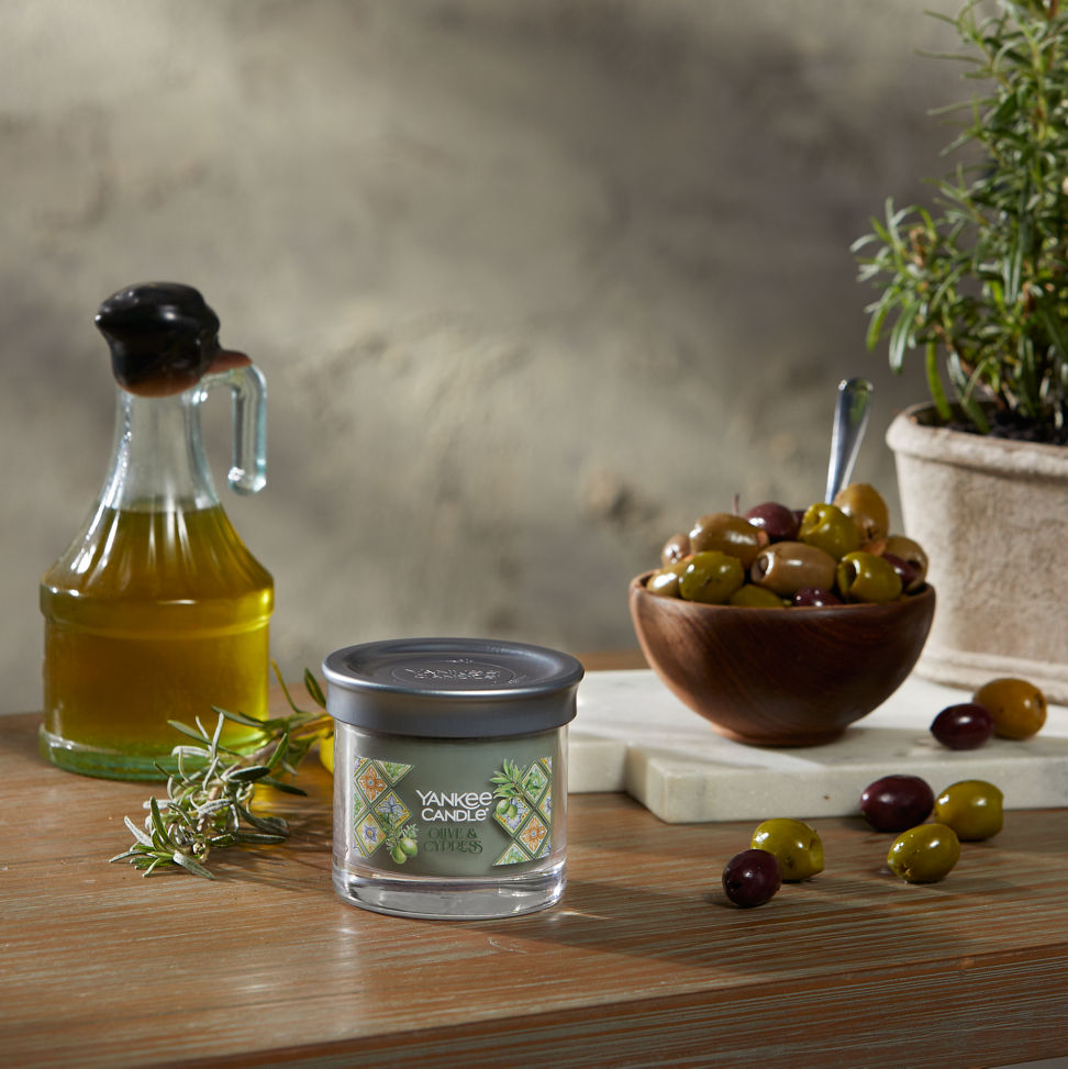 Hello, Italy! Collection Olive & Cypress Signature Small Tumbler Candle on a wood table with a jar of olive oil and a bowl of olives