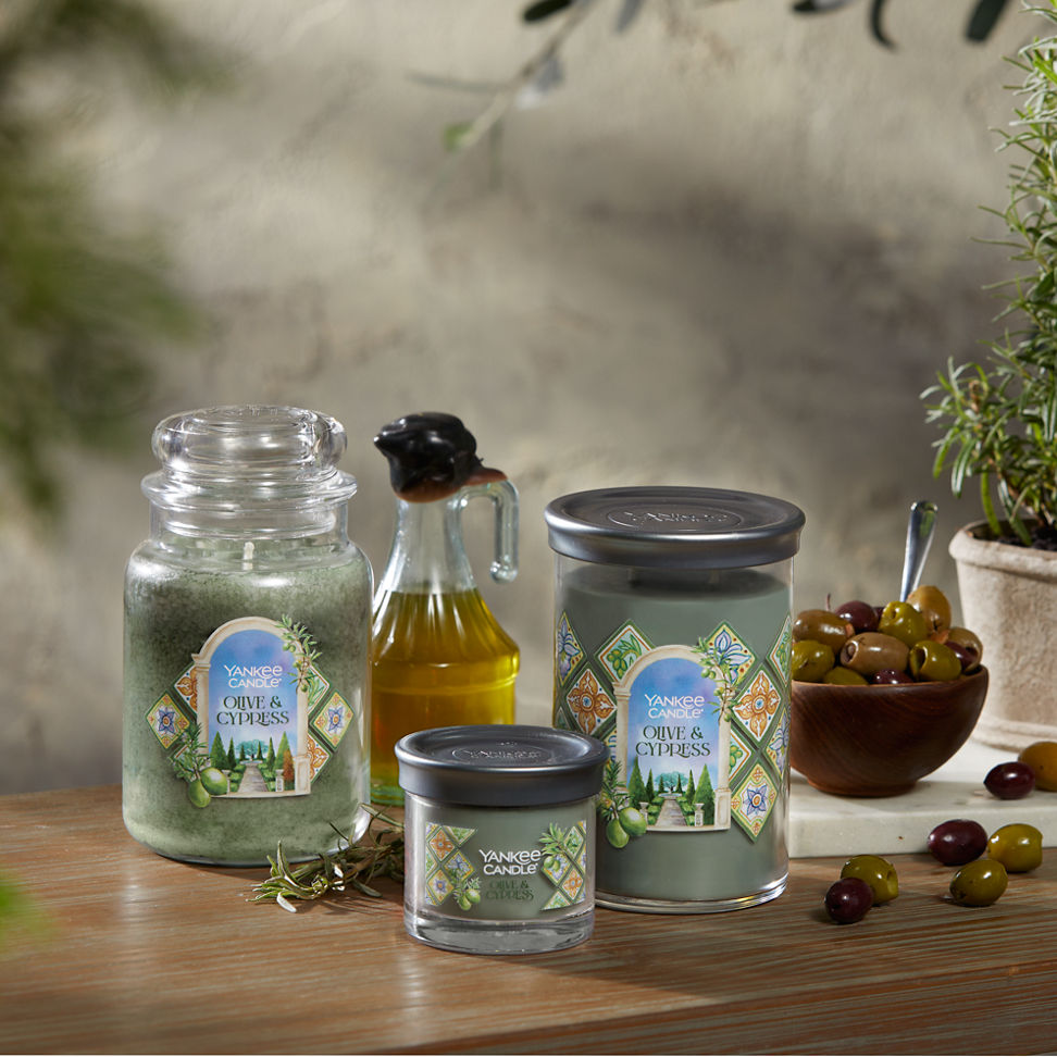 Hello, Italy! Collection Olive & Cypress Original Large Jar Candle, an Olive & Cypress Signature Small Tumbler Candle, and an Olive & Cypress Signature Large Tumbler Candle on a table with a jar of olive oil and a bowl of olives