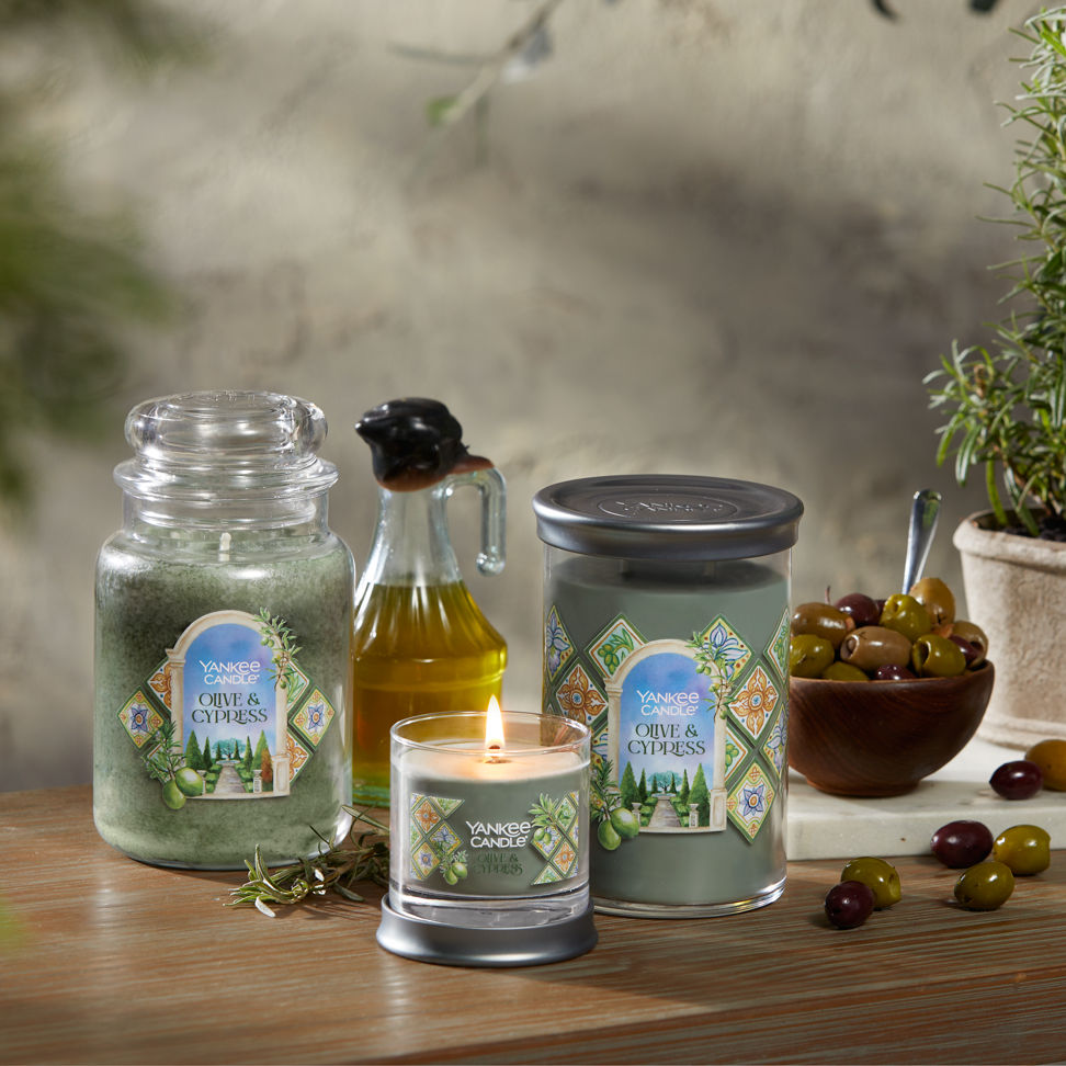 Hello, Italy! Collection Olive & Cypress Original Large Jar Candle, Olive & Cypress Signature Small Tumbler Candle, and an Olive & Cypress Signature Large Tumbler Candle on a table with a jar of olive oil and a bowl of olives