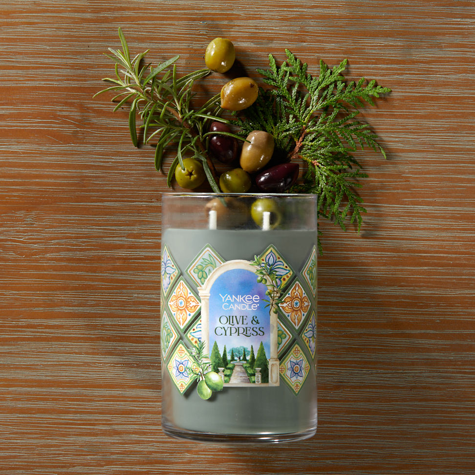 Hello, Italy! Collection Olive & Cypress Signature Large Tumbler Candle laying on a table with olives and sprigs of green leaves coming from the top of the candle