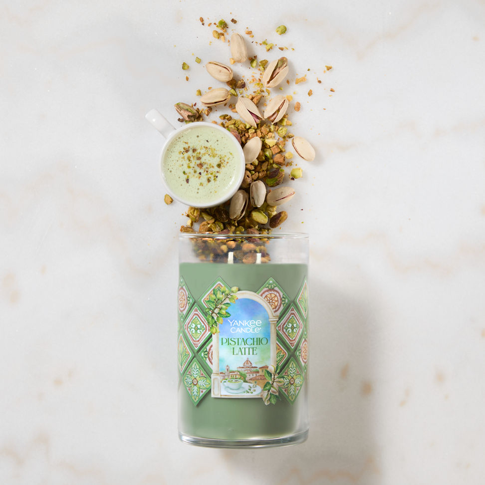 Hello, Italy! Collection Pistachio Latte Signature Large Tumbler Candle laying on a table with a latte and pistachios coming from the top of the candle