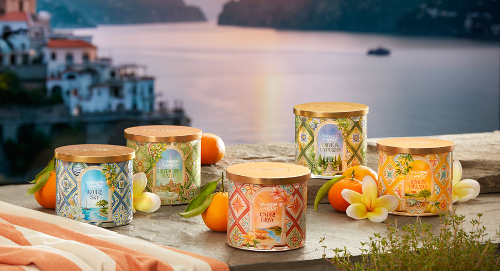 Hello, Italy! Collection Azure Sky 3-Wick Candle, Pistachio Latte 3-Wick Candle, Capri Glow 3-Wick Candle, Olive & Cypress 3-Wick Candle, and Lemon Gelato 3-Wick Candle on a rock surface surrounded by fruit and flowers overlooking water and a sunset